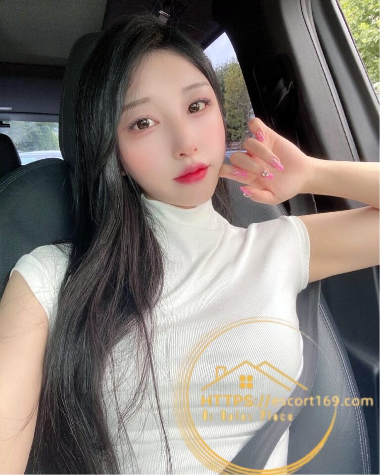 Selfie of a woman with long black hair in a white turtleneck top inside a car, hand near her face with pink nails and subtle makeup.