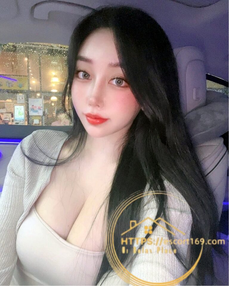 Self-portrait of a woman with long dark hair in a car, wearing a beige top, with soft makeup and nighttime city lights outside the window.