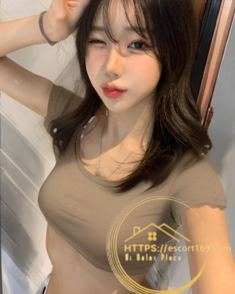Selfie of a young woman winking, with long brown hair, wearing a beige crop top, indoors; gold watermark overlay.